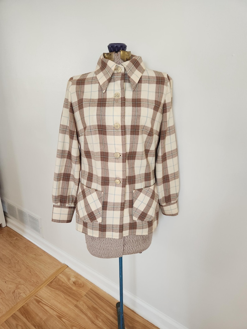 May include: A vintage plaid jacket in cream, brown, and light blue. The jacket features a collared neckline, button-down front, two front pockets, and long sleeves. The jacket is displayed on a mannequin.