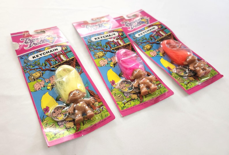 Puede incluir: Tres llaveros vintage de Troll Dolls en su embalaje original. Cada llavero presenta un mu&ntilde;eco troll marr&oacute;n con pelo colorido: amarillo, rosa y naranja. El embalaje es rosa y azul con las palabras "Troll Dolls" y "Keychain".