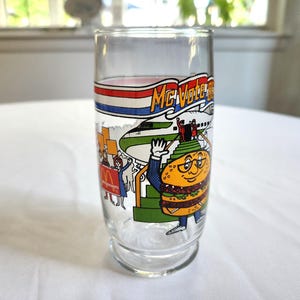 May include: A vintage glass McDonald's McVote Burger King glass with a cartoon burger character wearing a suit and glasses. The glass has a red, white, and blue banner with the text "McVote Burger King" and a cartoon airplane in the background.