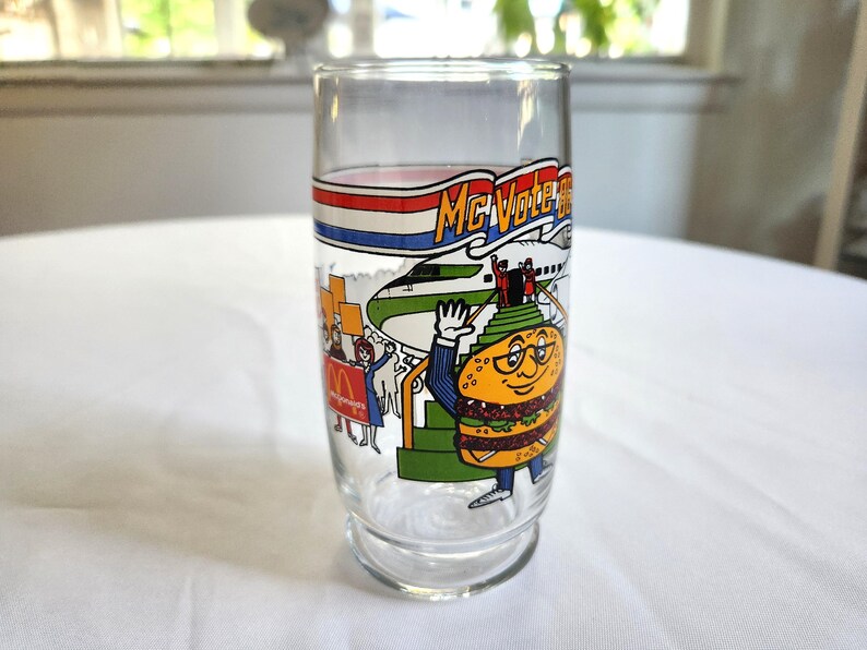 May include: A vintage glass McDonald's McVote Burger King glass with a cartoon burger character wearing a suit and glasses. The glass has a red, white, and blue banner with the text "McVote Burger King" and a cartoon airplane in the background.