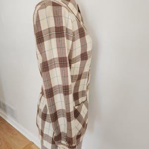 May include: A long-sleeved plaid shirt in cream, brown, and light blue. The shirt has a collared neckline, buttoned cuffs, and a single pocket. The fabric has a classic plaid pattern.