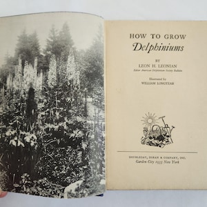 May include: An open book with a black and white photograph of a garden on the left page and text on the right. The book's title is "HOW TO GROW Delphiniums" by Leon H. Leonian, illustrated by William Longyear, published in 1935.