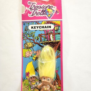 Puede incluir: Un llavero vintage de Treasure Trolls. El llavero presenta una peque&ntilde;a mu&ntilde;eca troll marr&oacute;n con cabello amarillo, adjunta a una tarjeta rosa y azul. La tarjeta tiene las palabras "KEYCHAIN" y "Treasure Trolls" impresas, junto con gr&aacute;ficos de dibujos animados.