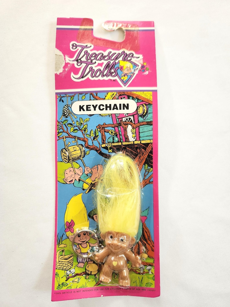 Puede incluir: Un llavero vintage de Treasure Trolls. El llavero presenta una peque&ntilde;a mu&ntilde;eca troll marr&oacute;n con cabello amarillo, adjunta a una tarjeta rosa y azul. La tarjeta tiene las palabras "KEYCHAIN" y "Treasure Trolls" impresas, junto con gr&aacute;ficos de dibujos animados.