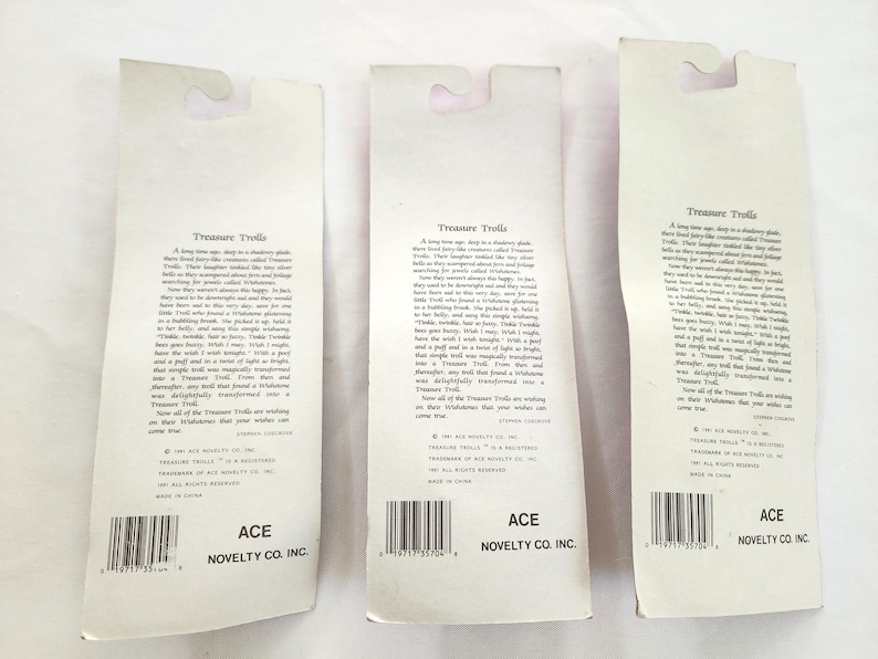 Puede incluir: Tres tarjetas de embalaje de juguetes vintage de Treasure Trolls. Cada tarjeta es blanca con una parte superior curvada y un c&oacute;digo de barras en la parte inferior. El texto "Treasure Trolls" est&aacute; impreso en la parte superior, con texto adicional debajo. La parte inferior de cada tarjeta dice "ACE NOVELTY CO. INC."