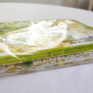 May include: A folded, packaged fitted sheet with a floral pattern in yellow and green. The sheet is in a clear plastic package with green trim. The package text reads "BOTTOM FITTED PERCALE SHEET (FITS 39 x 76 IN. MATTRESS)".