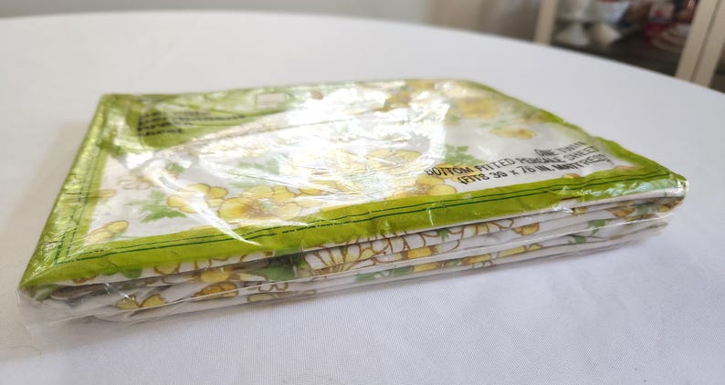 May include: A folded, packaged fitted sheet with a floral pattern in yellow and green. The sheet is in a clear plastic package with green trim. The package text reads "BOTTOM FITTED PERCALE SHEET (FITS 39 x 76 IN. MATTRESS)".