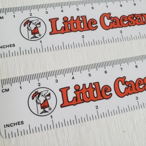 Puede incluir: Dos reglas de pl&aacute;stico blancas con un logotipo de Little Caesars rojo y blanco y una mascota de dibujos animados. Las reglas tienen marcas en pulgadas y cent&iacute;metros.