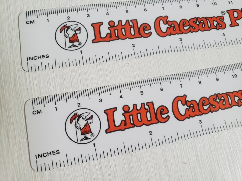 Puede incluir: Dos reglas de pl&aacute;stico blancas con un logotipo de Little Caesars rojo y blanco y una mascota de dibujos animados. Las reglas tienen marcas en pulgadas y cent&iacute;metros.