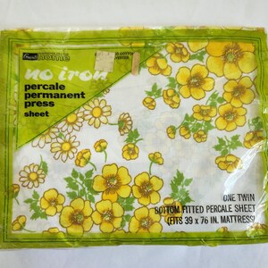 May include: A packaged twin-size percale sheet with a floral pattern. The sheet features yellow and white flowers with green leaves on a white background. The packaging is green with text that reads "no iron" and "bottom fitted percale sheet (fits 39 x 76 in. mattress)."