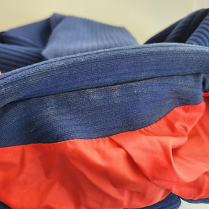 May include: A blue and red jacket with a ribbed collar and a red lining. The jacket is folded in half, showing the inside lining.