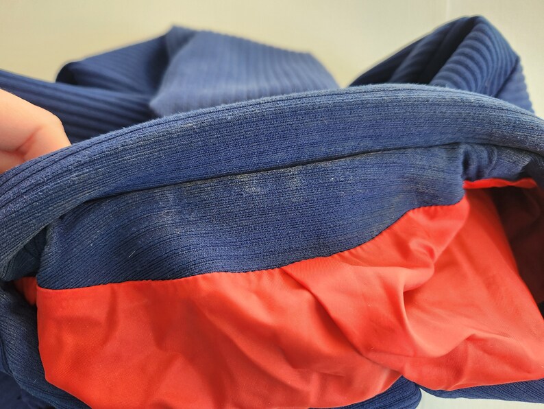 May include: A blue and red jacket with a ribbed collar and a red lining. The jacket is folded in half, showing the inside lining.