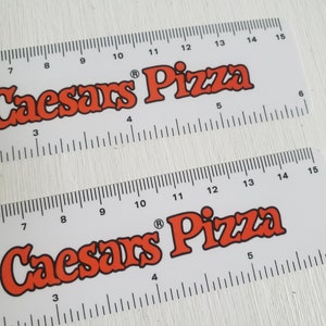 Puede incluir: Dos reglas blancas con marcas negras y las palabras "Little Caesars Pizza" en letras rojas. Las reglas miden 38 cm de largo.