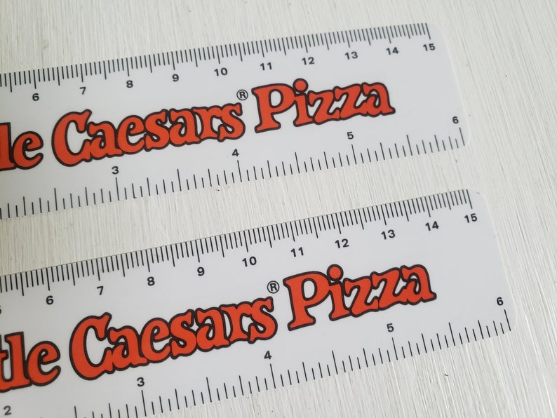 Puede incluir: Dos reglas blancas con marcas negras y las palabras "Little Caesars Pizza" en letras rojas. Las reglas miden 38 cm de largo.
