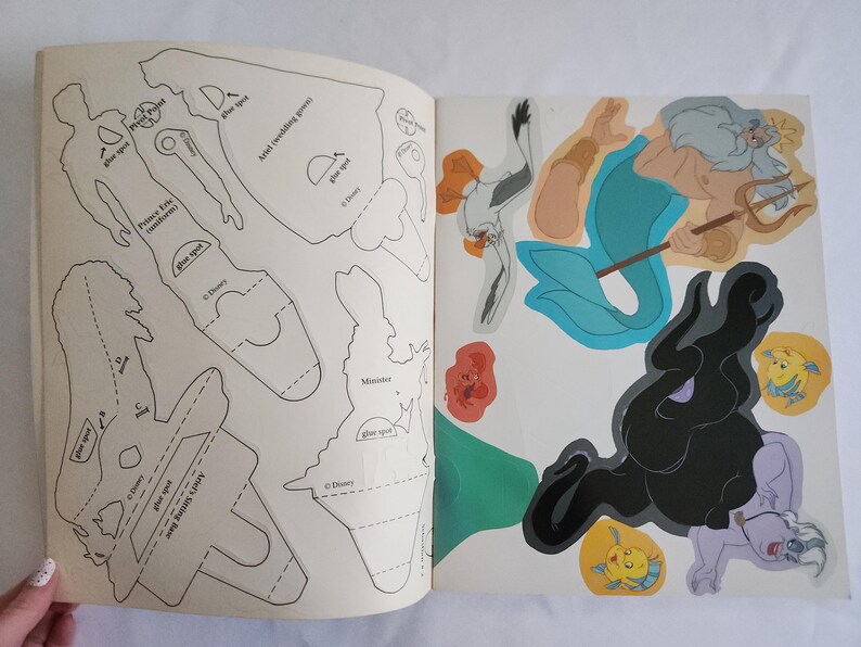 Puede incluir: Un libro de manualidades de Disney con contornos en blanco y negro para recortar figuras del Pr&iacute;ncipe Eric y Ariel en la p&aacute;gina izquierda, e ilustraciones a color del Rey Trit&oacute;n, &Uacute;rsula, Flounder y Sebasti&aacute;n en la p&aacute;gina derecha.