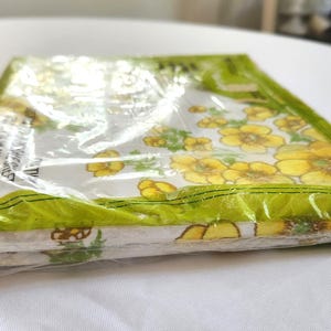 May include: A package of floral-patterned fabric napkins. The napkins are white with yellow flowers and green leaves. The package is sealed in clear plastic with a green border.