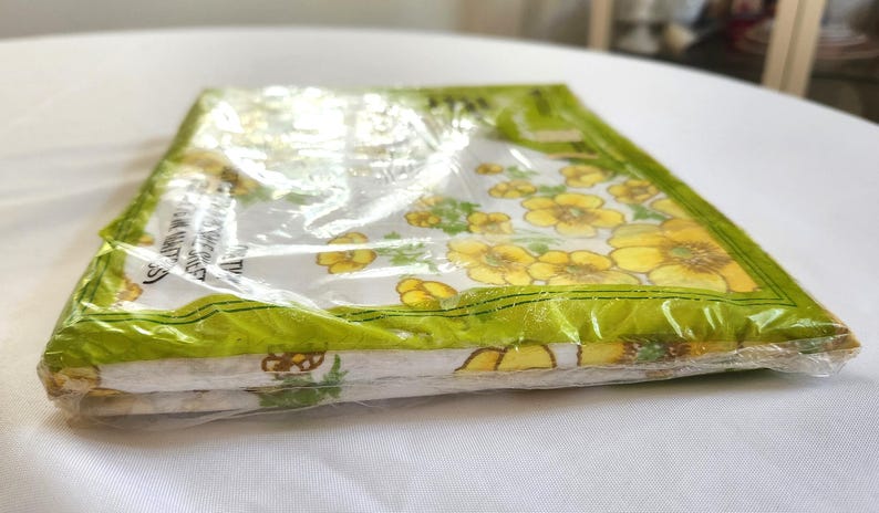 May include: A package of floral-patterned fabric napkins. The napkins are white with yellow flowers and green leaves. The package is sealed in clear plastic with a green border.