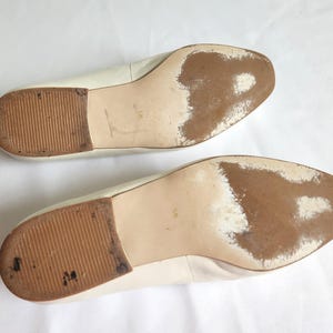 Puede incluir: Un par de bailarinas de cuero blanco roto, vistas desde las suelas. Las suelas muestran desgaste con decoloraci&oacute;n marr&oacute;n y tacones de goma texturizados. Los zapatos est&aacute;n sobre un fondo blanco.