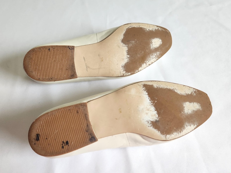 Puede incluir: Un par de bailarinas de cuero blanco roto, vistas desde las suelas. Las suelas muestran desgaste con decoloraci&oacute;n marr&oacute;n y tacones de goma texturizados. Los zapatos est&aacute;n sobre un fondo blanco.