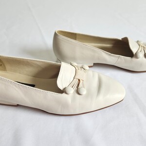 Puede incluir: Un par de mocasines de cuero blanco roto. Cada zapato presenta una puntera redondeada, un lazo decorativo con borlas y un tac&oacute;n bajo. Los zapatos est&aacute;n sobre un fondo blanco, mostrando su dise&ntilde;o cl&aacute;sico y color neutro.