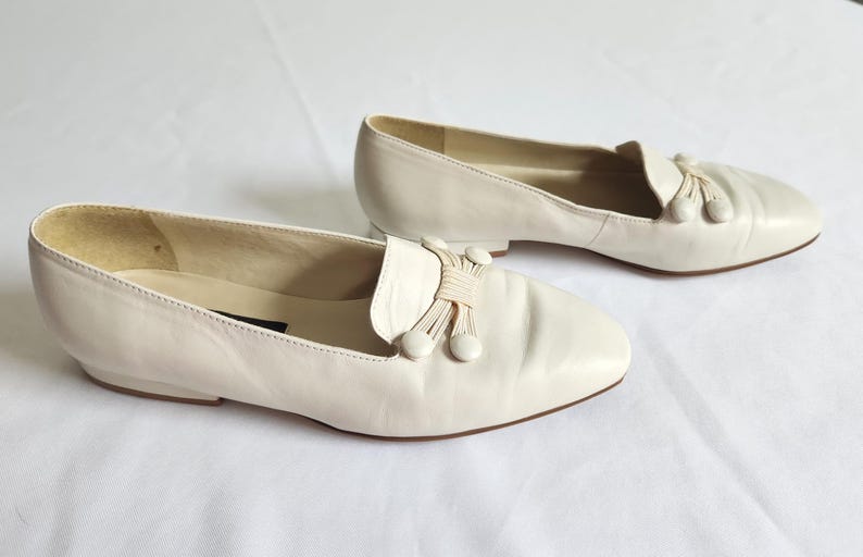 Puede incluir: Un par de mocasines de cuero blanco roto. Cada zapato presenta una puntera redondeada, un lazo decorativo con borlas y un tac&oacute;n bajo. Los zapatos est&aacute;n sobre un fondo blanco, mostrando su dise&ntilde;o cl&aacute;sico y color neutro.