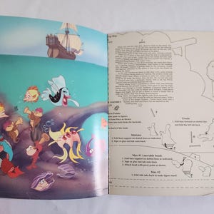 Puede incluir: Un libro abierto que presenta una ilustraci&oacute;n colorida de los personajes de La Sirenita bajo el agua en la p&aacute;gina izquierda e instrucciones de montaje en blanco y negro con texto en la p&aacute;gina derecha. La escena incluye un barco y peces.
