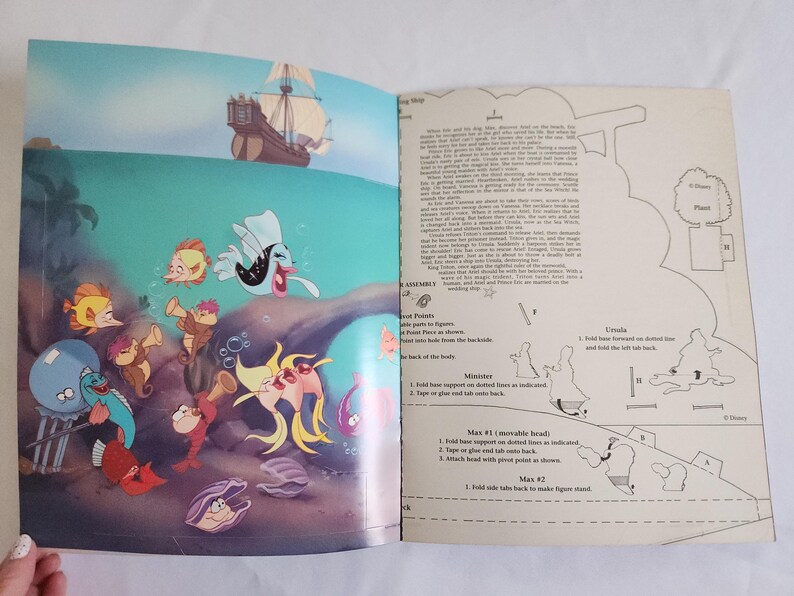 Puede incluir: Un libro abierto que presenta una ilustraci&oacute;n colorida de los personajes de La Sirenita bajo el agua en la p&aacute;gina izquierda e instrucciones de montaje en blanco y negro con texto en la p&aacute;gina derecha. La escena incluye un barco y peces.