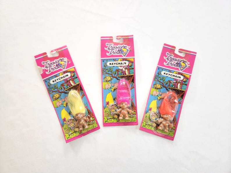 Puede incluir: Tres llaveros de Treasure Trolls en embalaje rosa. Cada llavero presenta una peque&ntilde;a mu&ntilde;eca troll con diferentes colores de pelo: amarillo, rosa y naranja. El embalaje incluye el texto "Treasure Trolls" y "KEYCHAIN".