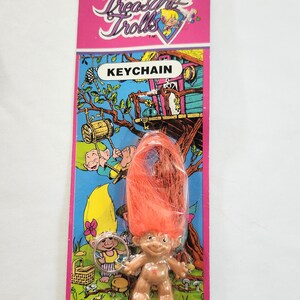 Puede incluir: Un llavero vintage de Treasure Trolls en una tarjeta rosa y verde. El llavero presenta una peque&ntilde;a figura de troll con cabello naranja. La tarjeta tiene las palabras "Treasure Trolls" y "KEYCHAIN" impresas.