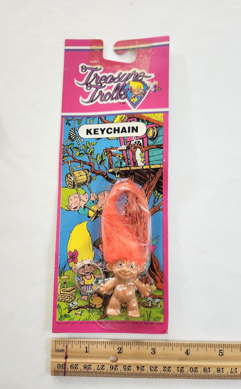 Puede incluir: Un llavero vintage de Treasure Trolls en una tarjeta rosa y verde. El llavero presenta una peque&ntilde;a figura de troll con cabello naranja. La tarjeta tiene las palabras "Treasure Trolls" y "KEYCHAIN" impresas.
