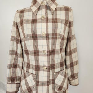 May include: A vintage button-down jacket in a cream, brown, and light blue plaid pattern. The jacket features a collared neckline, long sleeves, two front pockets, and four cream-colored buttons. The jacket is displayed on a mannequin.