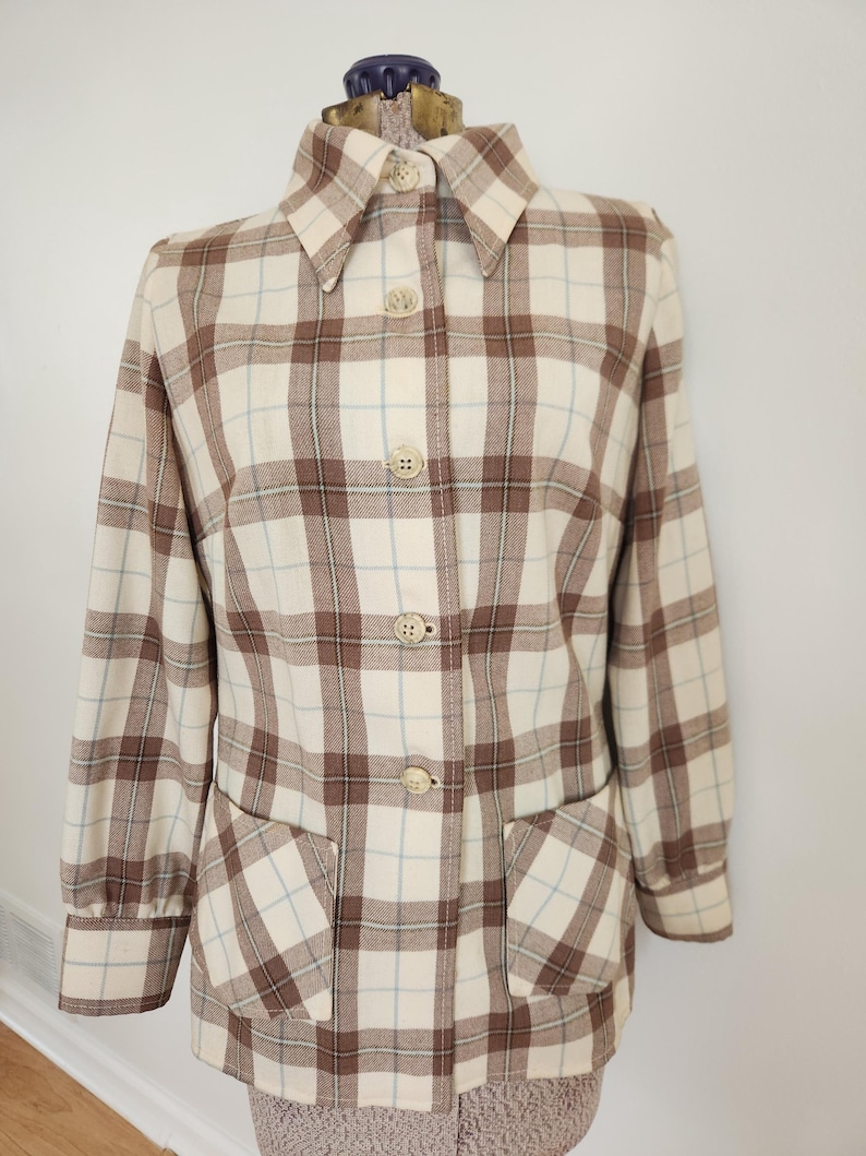 May include: A vintage button-down jacket in a cream, brown, and light blue plaid pattern. The jacket features a collared neckline, long sleeves, two front pockets, and four cream-colored buttons. The jacket is displayed on a mannequin.