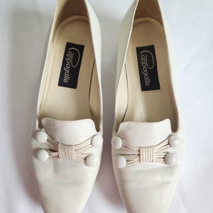 Puede incluir: Un par de mocasines de cuero blanco roto con lazo y detalles de botones. Los zapatos tienen una puntera redondeada y un tac&oacute;n peque&ntilde;o. La marca "cappagallo" est&aacute; impresa en una etiqueta negra en el interior.