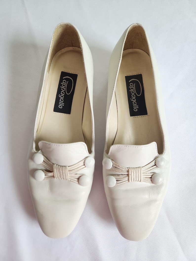 Puede incluir: Un par de mocasines de cuero blanco roto con lazo y detalles de botones. Los zapatos tienen una puntera redondeada y un tac&oacute;n peque&ntilde;o. La marca "cappagallo" est&aacute; impresa en una etiqueta negra en el interior.
