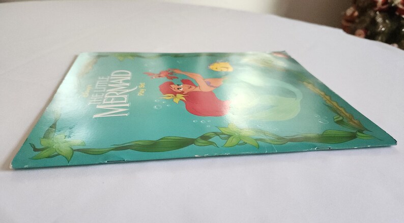 Puede incluir: Un paquete plano y rectangular de Disney The Little Mermaid Play Set con un fondo verde azulado e ilustraciones de algas verdes. El frente muestra una sirena de cabello rojo, un peque&ntilde;o pez amarillo y el texto The Little Mermaid Play Set.