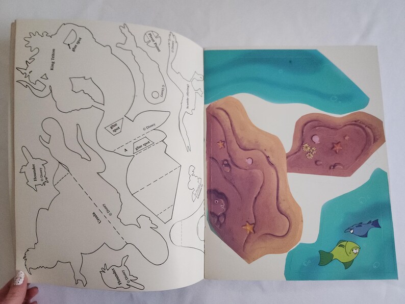 Puede incluir: Un libro de actividades abierto que muestra contornos en blanco y negro de personajes de Disney, incluidos el Rey Trit&oacute;n, &Uacute;rsula y Flounder en la p&aacute;gina izquierda, con formas recortables de colores de terreno submarino y peces a la derecha.
