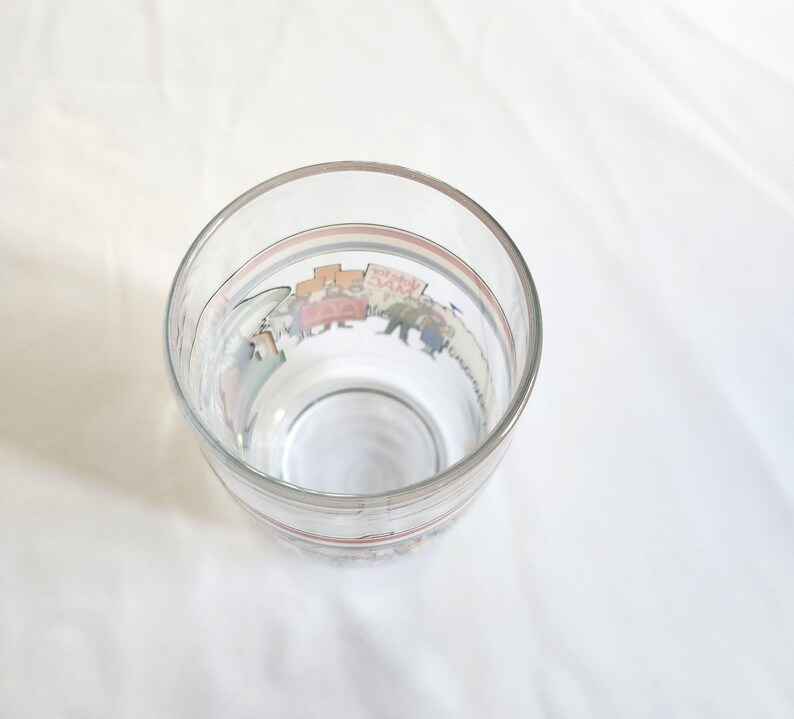 May include: A clear glass drinking glass with a colorful cartoon illustration of people marching around the rim.