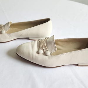 Puede incluir: Un par de mocasines de cuero color crema. Cada zapato presenta una borla decorativa y dos peque&ntilde;os detalles redondos. Los zapatos tienen un tac&oacute;n bajo y una puntera redondeada. Los mocasines se muestran sobre una superficie blanca.