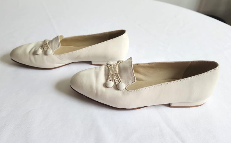 Puede incluir: Un par de mocasines de cuero color crema. Cada zapato presenta una borla decorativa y dos peque&ntilde;os detalles redondos. Los zapatos tienen un tac&oacute;n bajo y una puntera redondeada. Los mocasines se muestran sobre una superficie blanca.