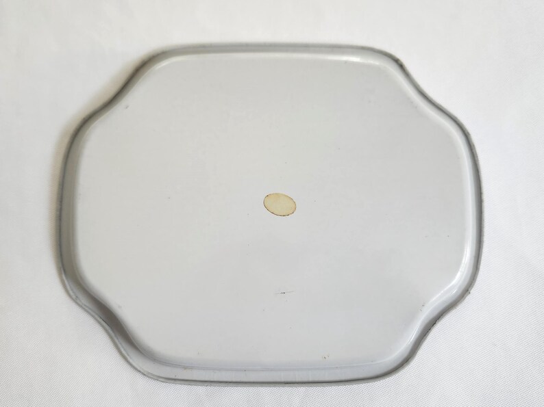 Op de afbeelding: Een vintage, lichtgrijs metalen dienblad met een geschulpte rand. De lade heeft een iets verhoogde rand en een kleine, ovale sticker in het midden. De vorm van de lade is een aangepaste achthoek.