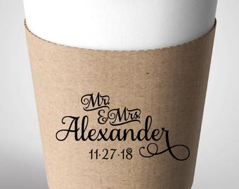 May include: A white paper coffee cup with a black lid and a brown paper sleeve. The sleeve has the text "Mr & Mrs Alexander 11.27.18" in black script. The cup is designed for hot beverages.