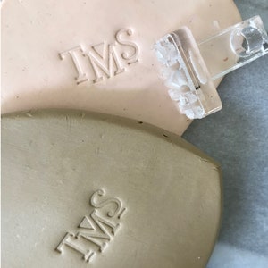 May include: Two close-up images of a light brown and a beige clay surface with the letters "TMS" embossed into the surface. A clear plastic stamp with the letters "TMS" is shown on top of the light brown clay surface.