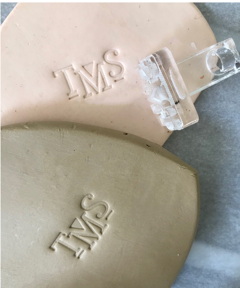 May include: Two close-up images of a light brown and a beige clay surface with the letters "TMS" embossed into the surface. A clear plastic stamp with the letters "TMS" is shown on top of the light brown clay surface.