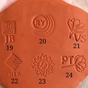 May include: Close-up of a terracotta clay tile with six different stamp impressions. The stamps are numbered 19 through 24. The stamps feature various designs, including geometric shapes, initials, and floral motifs.