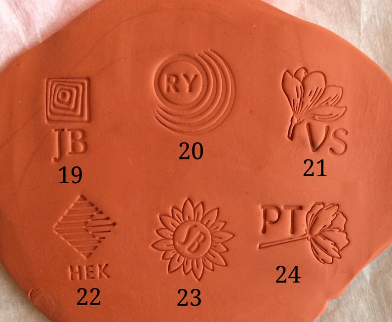 May include: Close-up of a terracotta clay tile with six different stamp impressions. The stamps are numbered 19 through 24. The stamps feature various designs, including geometric shapes, initials, and floral motifs.