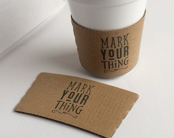 May include: A brown cardboard coffee cup sleeve with the text "Mark Your Thing" printed in black.