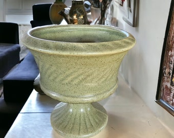 May include: A green ceramic planter with a speckled finish and a fluted base. The planter is sitting on a wooden table.