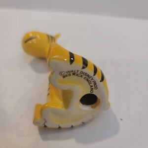May include: A yellow and black ceramic figurine of Tigger from Winnie the Pooh. The figurine is sitting on its hind legs with its tail curled up. The bottom of the figurine has the text "Walt Disney Prod. Beswick England".