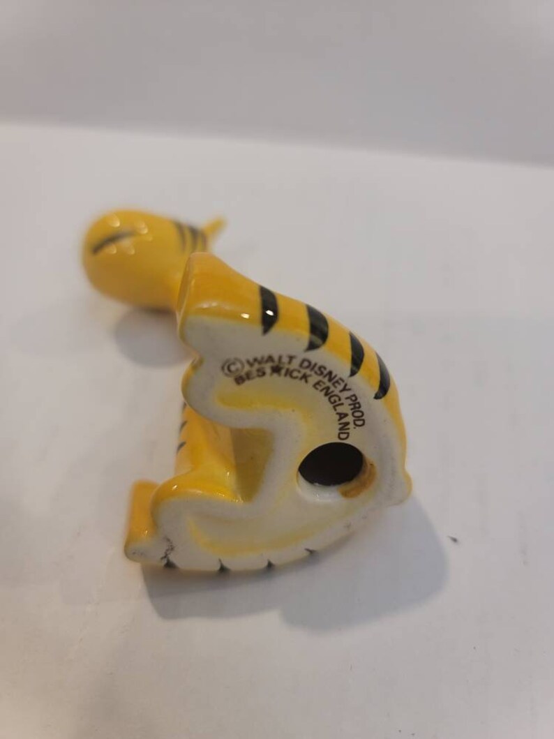 May include: A yellow and black ceramic figurine of Tigger from Winnie the Pooh. The figurine is sitting on its hind legs with its tail curled up. The bottom of the figurine has the text "Walt Disney Prod. Beswick England".