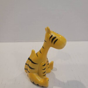 May include: A small ceramic figurine of a tiger sitting down. The tiger is yellow with black stripes and has a simple, cartoon-like design.